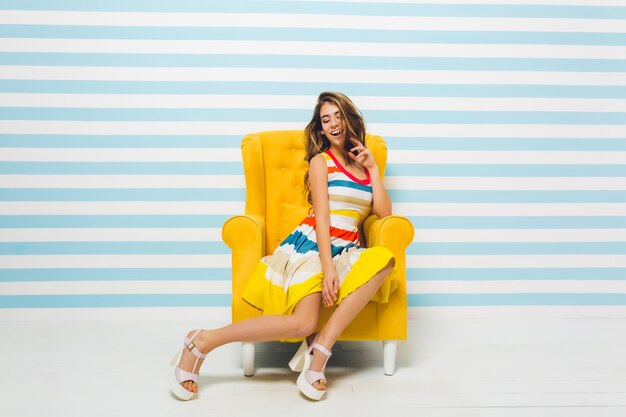 Mujer joven emocionada con hermosa sonrisa escalofriante en un cómodo sillón. Impresionante chica de pelo largo con vestido de moda y zapatos de tacón sentado en muebles amarillos con los ojos cerrados.