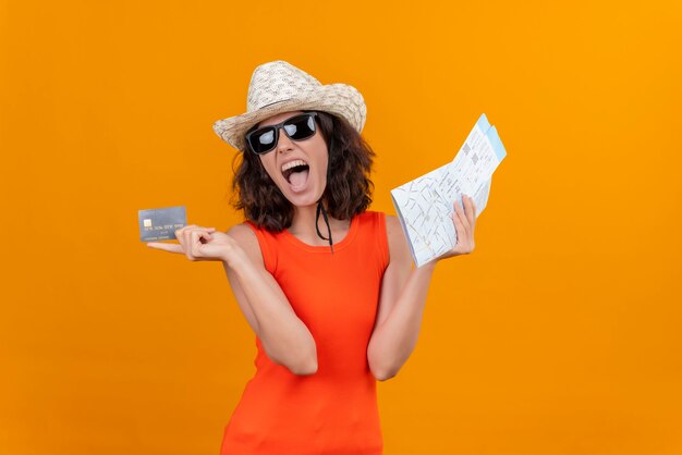 Una mujer joven emocionada con el pelo corto en una camisa naranja con sombrero para el sol y gafas de sol con mapa y tarjeta de crédito