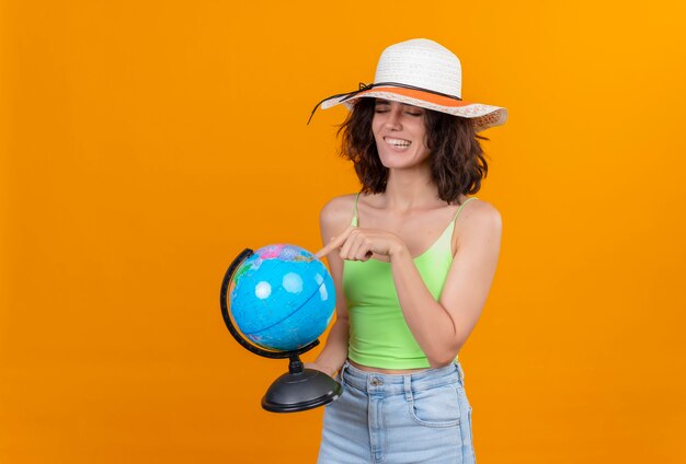 Una mujer joven emocionada con el pelo corto en la parte superior de la cosecha verde con sombrero para el sol apuntando a un globo con el dedo índice