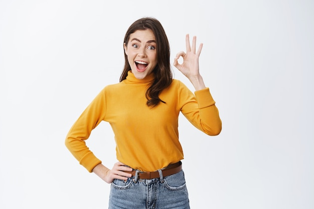 Mujer joven emocionada que anuncia un buen producto, que muestra el signo de OK y dice que sí, aprueba algo genial, elogia la elección increíble, de pie feliz contra la pared blanca