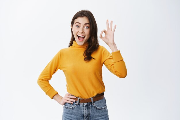 Mujer joven emocionada que anuncia un buen producto, que muestra el signo de OK y dice que sí, aprueba algo genial, elogia la elección increíble, de pie feliz contra la pared blanca