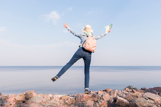 Foto gratuita mujer joven emocionada que se coloca encima de la roca que sostiene el mapa en la mano que pasa por alto el mar contra el cielo azul