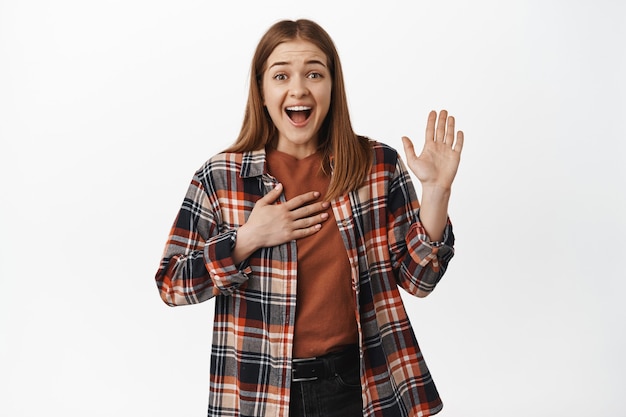 Foto gratuita mujer joven emocionada que levanta la mano y coloca la palma en el corazón, sonriendo feliz, se presenta, mi nombre es gesto, saluda, dice la verdad, hace una promesa, pared blanca.