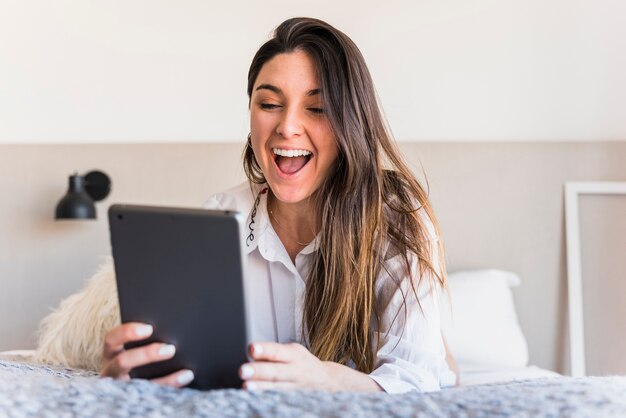 Mujer joven emocionada que miente en la alfombra usando la tableta digital