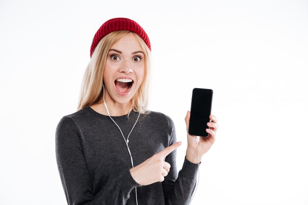 Mujer joven emocionada en sombrero que señala el dedo en el teléfono móvil de la pantalla en blanco