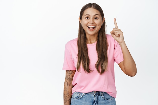 Mujer joven emocionada tiene una gran idea, apuntando con el dedo hacia arriba, gesto eureka, mostrando algo arriba en blanco