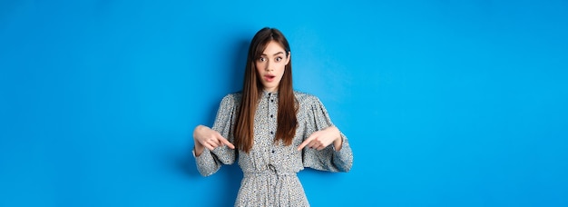 Mujer joven emocionada en vestido apuntando con el dedo hacia abajo en el logotipo mirando intrigado a la cámara de pie en b