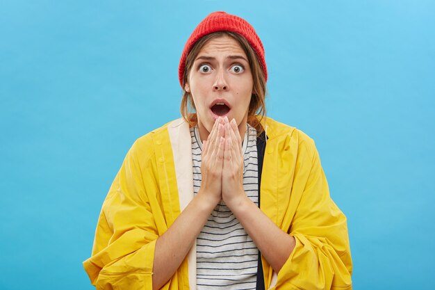 Mujer joven emocional asombrada en gabardina amarilla y sombrero rojo posando contra la pared azul en blanco con la mandíbula caída y los ojos abiertos, tomada por sorpresa. Actitud y reacción humana