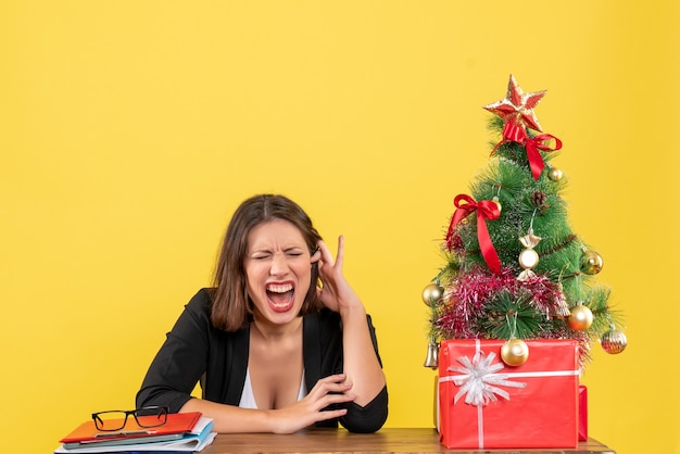 Mujer joven emocional cerrando los ojos sentado en una mesa cerca del árbol de Navidad decorado en la oficina en amarillo