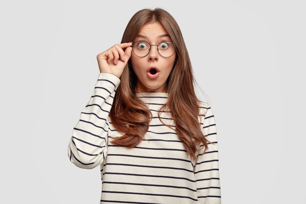 Mujer joven emocional con gafas posando contra la pared blanca