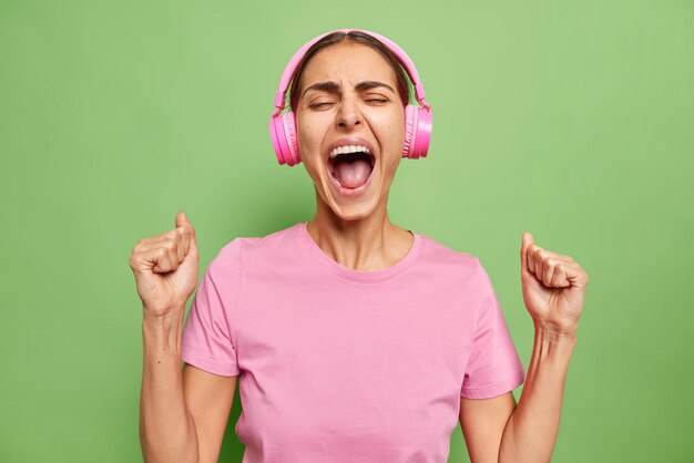 Mujer joven emocional grita con emoción aprieta los puños mantiene la boca abierta grita de éxito escucha música en auriculares usa camiseta rosa aislada sobre pared verde vivo