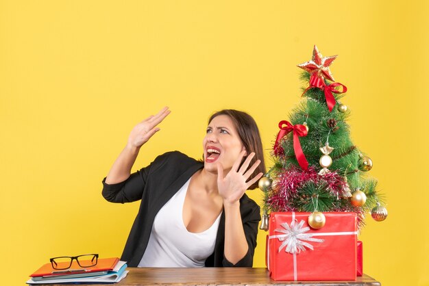 Mujer joven emocional mirando algo sentado en una mesa cerca del árbol de Navidad decorado en la oficina en amarillo