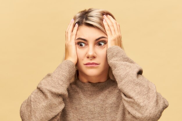 Mujer joven emocional en suéter acogedor de gran tamaño cogidos de la mano en la cabeza en pánico y conmoción, con mirada olvidadiza. Chica nerviosa frustrada que se asusta y se preocupa porque se equivocó