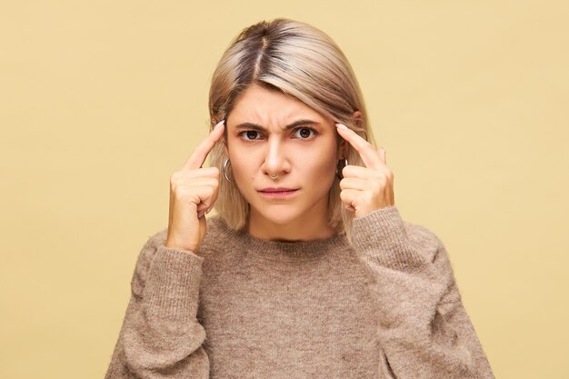 Mujer joven emocional en suéter que sufre de dolor de cabeza, tiene tensión cerebral masajeando las sienes con los dedos, sintiéndose frustrada por la indignación, frunciendo el ceño, tratando de pensar