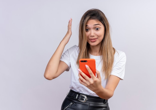 Una mujer joven encantadora emocionada en camiseta blanca levantando la mano mientras mira el teléfono móvil