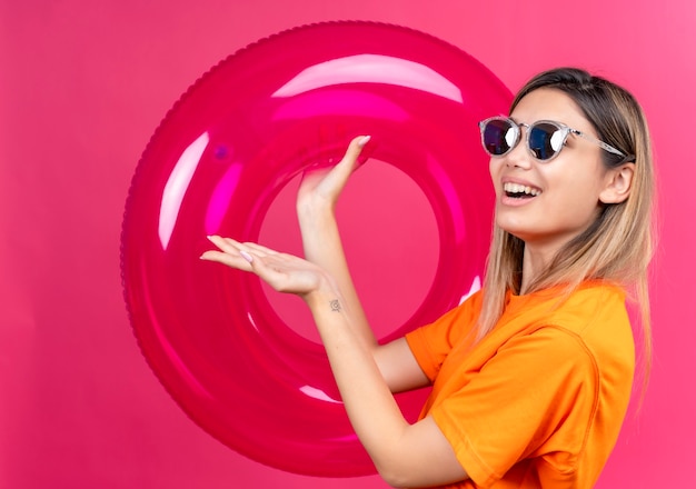 Una mujer joven encantadora feliz en una camiseta naranja con gafas de sol sonriendo y mirando mientras sostiene el anillo inflable rosa en una pared rosa