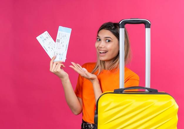 Una mujer joven encantadora feliz en una camiseta naranja que muestra boletos de avión con maleta amarilla en una pared rosa