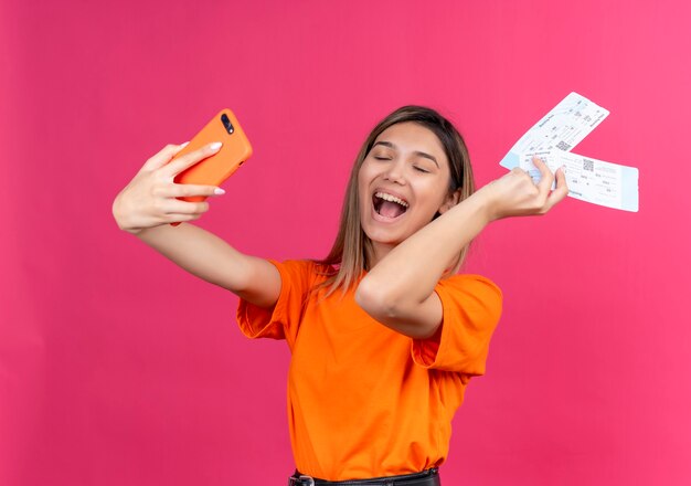 Una mujer joven encantadora feliz en una camiseta naranja sonriendo y tomando selfie con teléfono móvil mientras muestra boletos de avión en una pared rosa