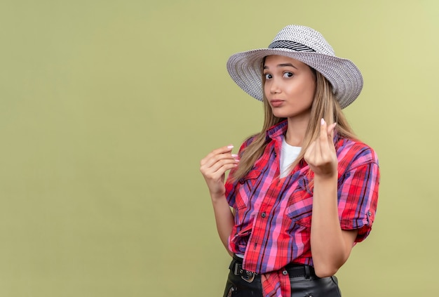 Una mujer joven encantadora molesta en una camisa a cuadros con sombrero que no muestra ningún gesto de dinero con la mano