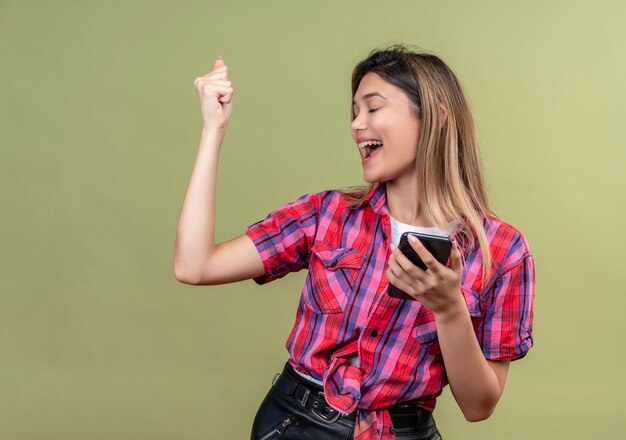 Una mujer joven encantadora muy feliz en una camisa a cuadros sosteniendo el teléfono móvil mientras levanta el puño cerrado