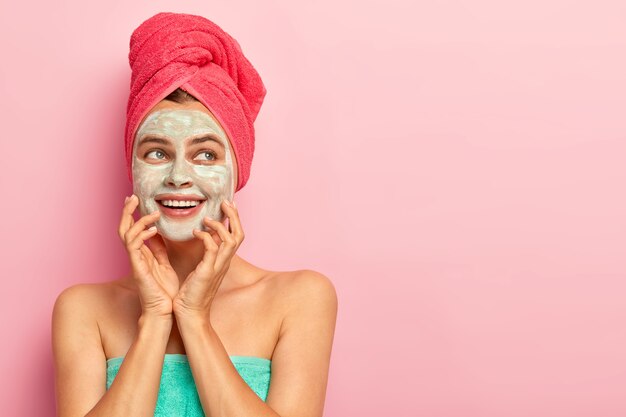 Mujer joven encantadora soñadora con máscara de arcilla, toca las mejillas, tiene expresión de alegría en la cara, dientes blancos, sueña con una piel perfecta, se ha envuelto una toalla en la cabeza, disfruta de tratamientos de belleza