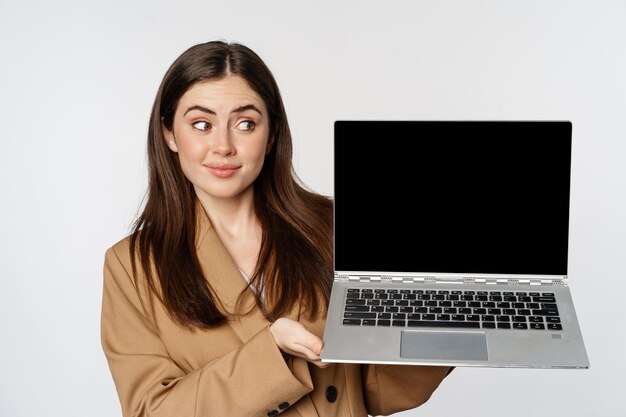 La mujer joven se encoge y se ve escéptica mostrando la pantalla del portátil de pie sobre fondo blanco.