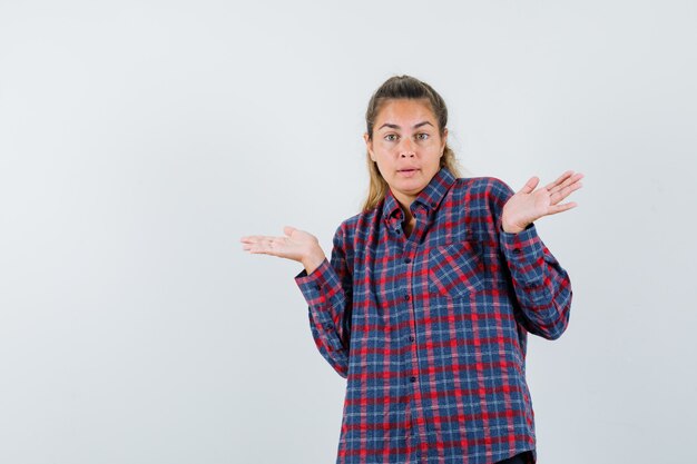 Mujer joven encogiéndose de hombros en camisa a cuadros y mirando perplejo