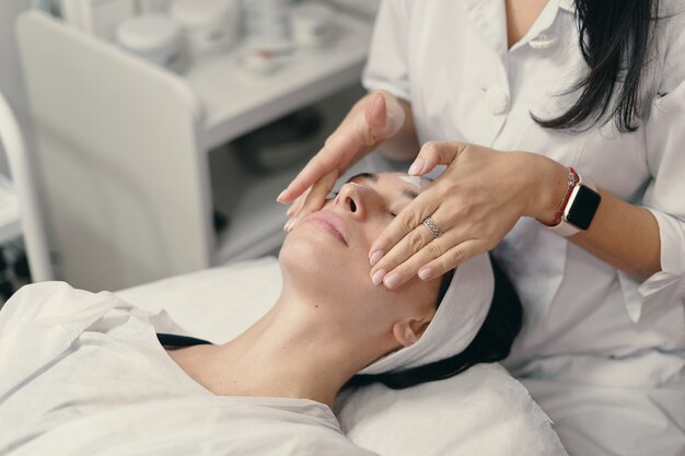 Mujer joven se encuentra con los ojos cerrados, cosmetóloga haciendo procedimiento
