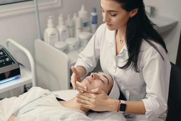 Mujer joven se encuentra con los ojos cerrados, cosmetóloga haciendo procedimiento