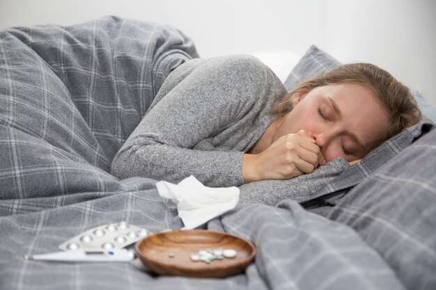 Mujer joven enferma acostada en la cama con los ojos cerrados, tosiendo