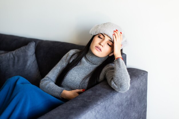 Mujer joven enferma en homewear gris que se sienta en cama con la computadora portátil que trabaja, mirando el termómetro después de medir temperatura. Enfermedad, concepto independiente.