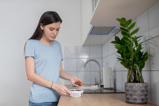 Mujer joven enfocada en calma poniendo comida seca para perros en el cuenco de cerámica colocado sobre la encimera de la cocina