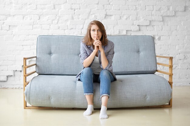 Mujer joven enfocada preocupada en jeans y calcetines blancos sentada en un sofá gris contra el fondo de la pared de ladrillo blanco, con mirada pensativa concentrada, frunciendo los labios, pensando en ideas de renovación del hogar
