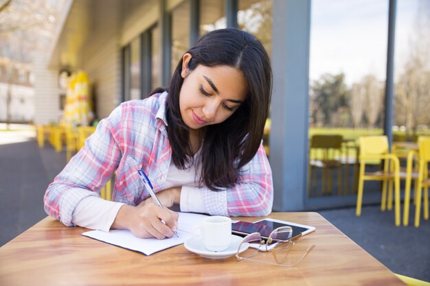 Mujer joven enfocada que hace notas en café al aire libre