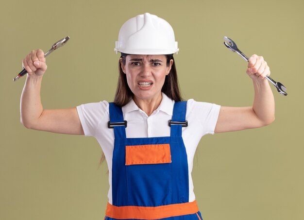 Mujer joven enojada del constructor en uniforme que levanta las llaves abiertas aisladas en la pared verde oliva