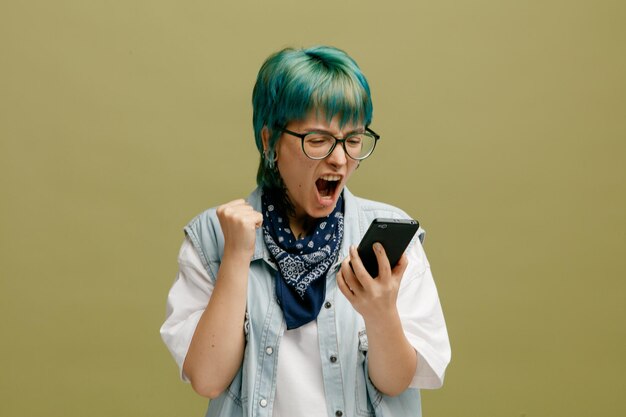 Mujer joven enojada con gafas bandana en el cuello sosteniendo un teléfono móvil mirándolo manteniendo el puño en el aire gritando aislado en un fondo verde oliva