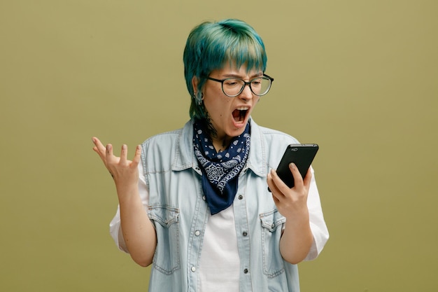 Mujer joven enojada con gafas bandana en el cuello sosteniendo un teléfono móvil mirándolo mostrando la mano vacía aislada en un fondo verde oliva