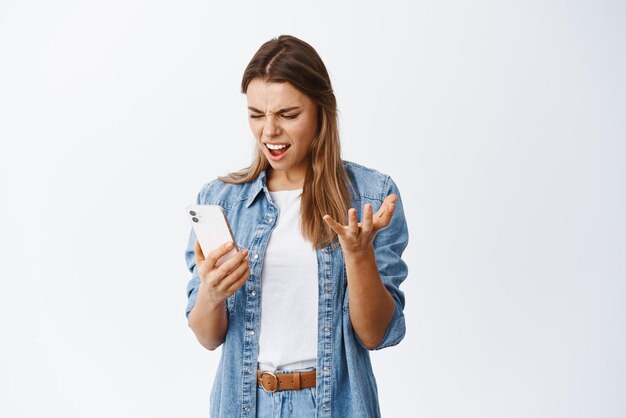 Mujer joven enojada y molesta mirando el teléfono móvil enojada quejándose de ver malas noticias en la pantalla del teléfono inteligente de pie contra el fondo blanco