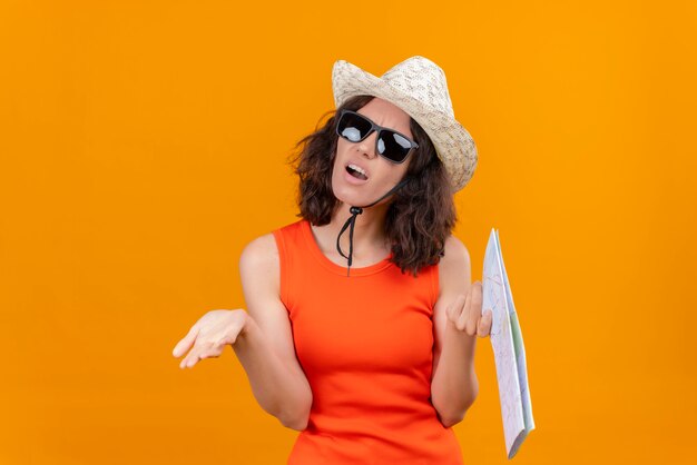 Una mujer joven enojada con el pelo corto en una camisa naranja con sombrero para el sol y gafas de sol sosteniendo el mapa abriendo las manos