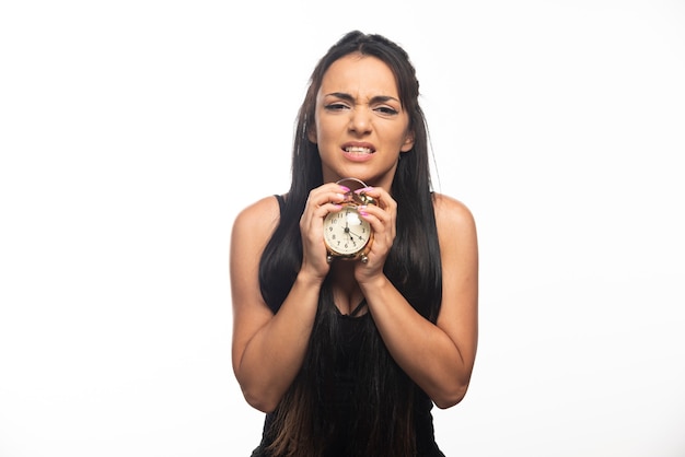 Mujer joven enojada sosteniendo un reloj de alarma en la pared blanca