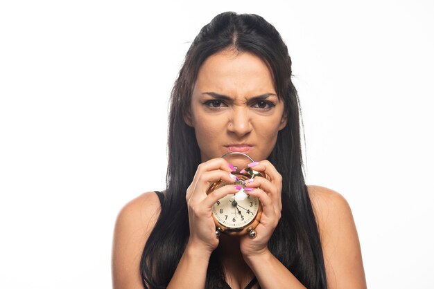 Mujer joven enojada sosteniendo un reloj de alarma en la pared blanca