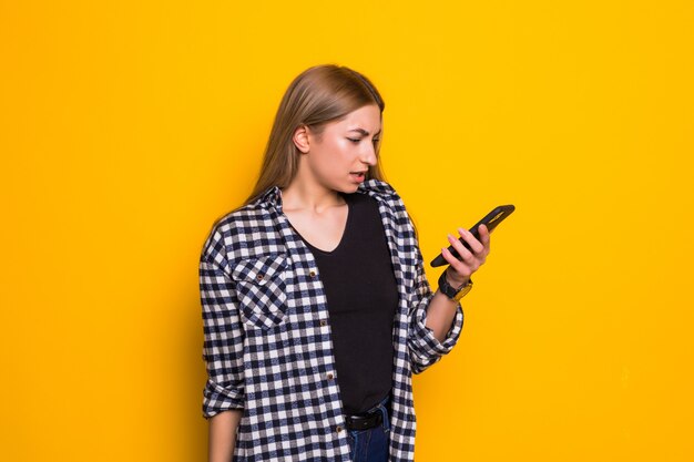 Mujer joven enojada con teléfono celular. Retrato de una mujer con un teléfono móvil, sobre una pared amarilla