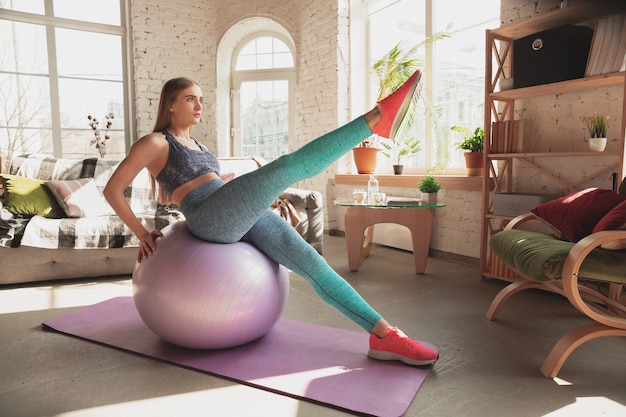 Mujer joven enseñando en casa cursos en línea de fitness, estilo de vida aeróbico y deportivo mientras está en cuarentena. Ponerse activo mientras está aislado, bienestar, concepto de movimiento. Ejercicios con fitball para la parte inferior del cuerpo.