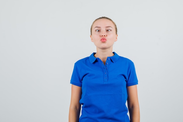 Mujer joven entrecerrando los ojos con los labios doblados en camiseta azul y mirando divertido