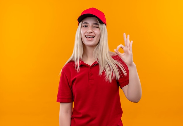 Mujer joven de entrega parpadeó vistiendo camiseta roja y gorra en soporte dental mostrando gesto okey en pared naranja aislada