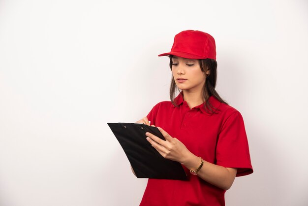 Mujer joven de entrega en uniforme rojo escribiendo en el portapapeles sobre fondo blanco.