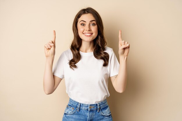 Mujer joven entusiasta, clienta señalando con el dedo hacia arriba y sonriendo, mostrando pancarta o logotipo, de pie contra un fondo beige.