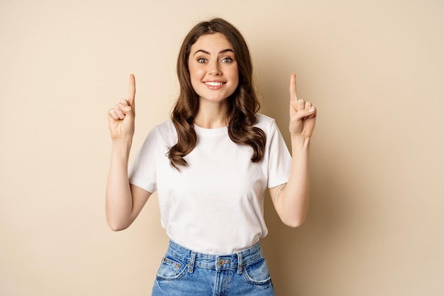 Mujer joven entusiasta, clienta señalando con el dedo hacia arriba y sonriendo, mostrando pancarta o logotipo, de pie contra un fondo beige.