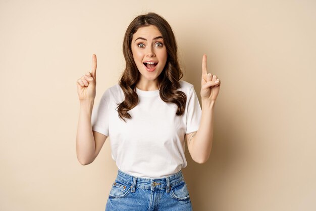 Mujer joven entusiasta, clienta señalando con el dedo hacia arriba y sonriendo, mostrando pancarta o logotipo, de pie contra un fondo beige