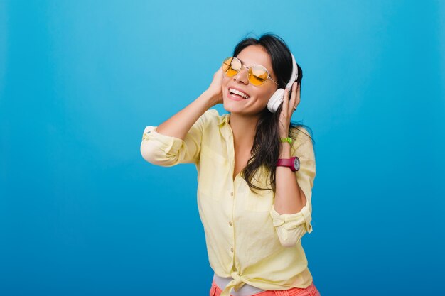 Mujer joven entusiasta en elegante camisa amarilla y pulsera rosa tocando auriculares mientras disfruta de la canción. Foto de interior de dichosa niña hispana con cabello castaño oscuro brillante posando.